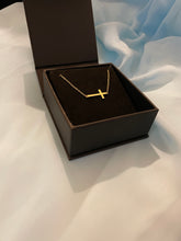 Load image into Gallery viewer, A gold sideways cross choker necklace inside a black jewelry box on a beautiful soft blue hued background. 
