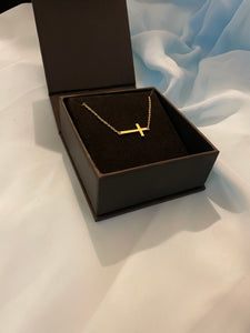 A gold sideways cross choker necklace inside a black jewelry box on a beautiful soft blue hued background. 